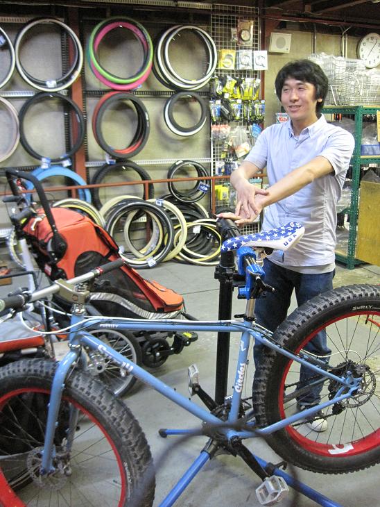 Left side view of a light blue Surly Pugsley fat bike on a stand, with a person standing behind it, inside a bike shop